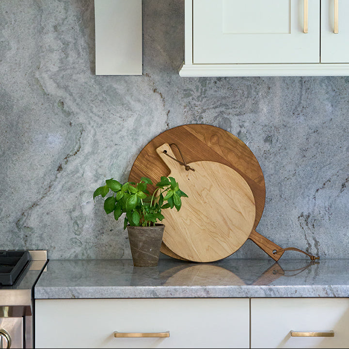 A close-up of a modern kitchen backsplash featuring a gray marble pattern, white cabinets with gold handles, a gold range hood trim, and two wooden cutting boards on a gray stone countertop next to a small potted green plant.