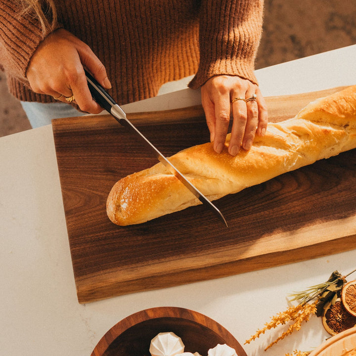 Person cutting a loaf of bread on a wooden cutting board with a knife.