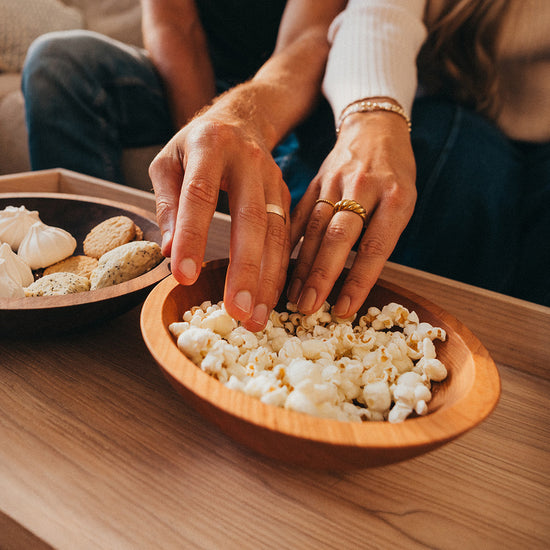 A close-up lifestyle shot of a person's hands reaching for popcorn in a round wooden bowl on a wooden table, with another bowl of snacks in the background.