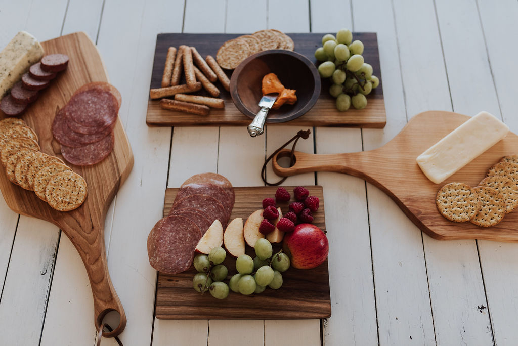 Bulk Gifts for employees come in many forms, like a collection of cutting and charcuterie boards. Four different styles of cutting boards by the Holland Bowl Mill, each wiith various fruits, vegetables, cheeses, and breads.