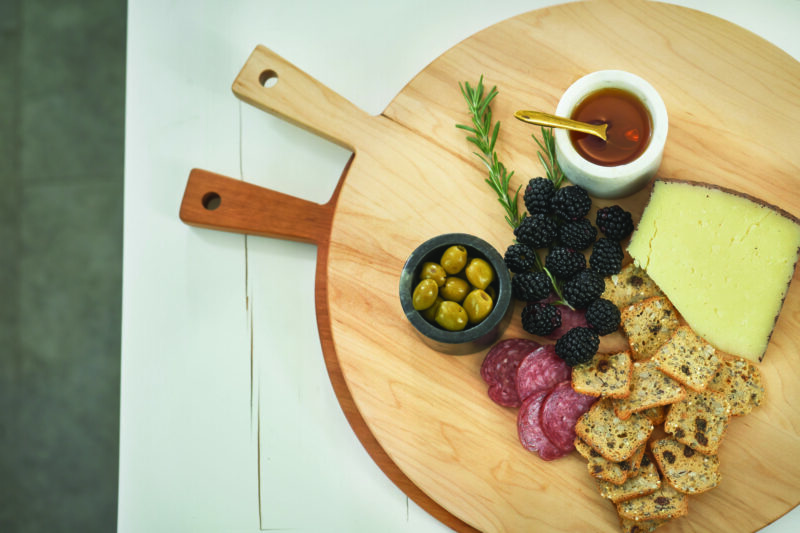 An over the top view of a round wooden serving bowl set with charcuterie setup: salami, crackers, berries and olives.