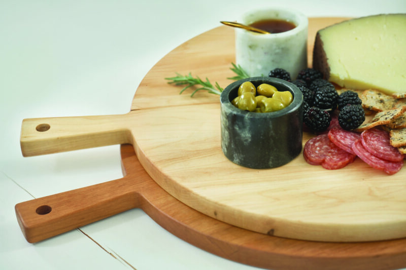 A close-up view of a round wooden serving bowl set with charcuterie setup: salami, crackers, berries and olives.