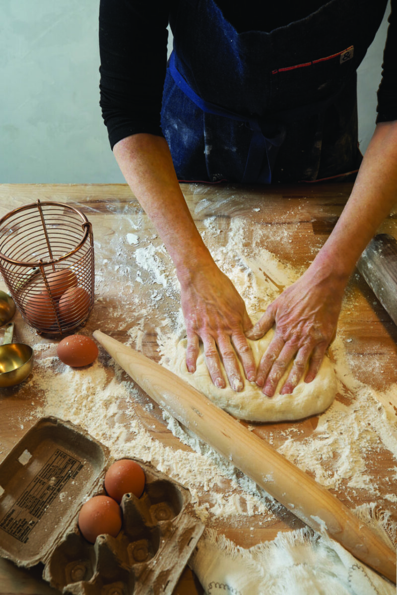 A baker uses a French rolling pin handy while kneading dough on a flour-topped kitchen counter.