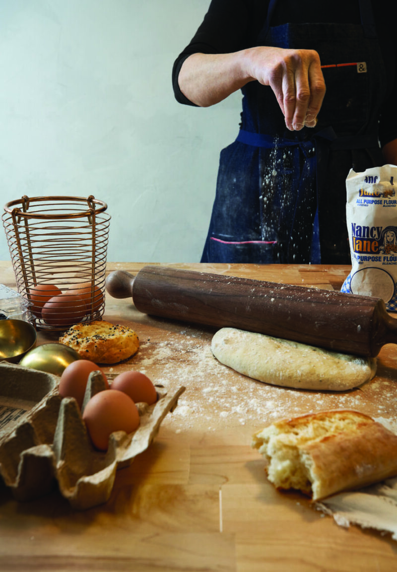 A woman sprinkles flour over a Shaker designed wooden rolling pin sits on top of a dough ball ready to be rolled into a crust.