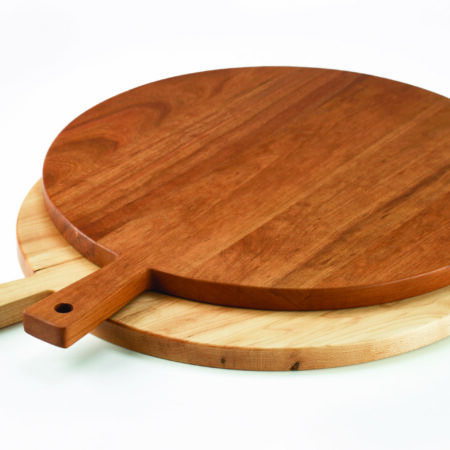 A Maple round wooden serving board lies on top of a Cherry round wooden serving bowl. Each has a small rectangular handle.