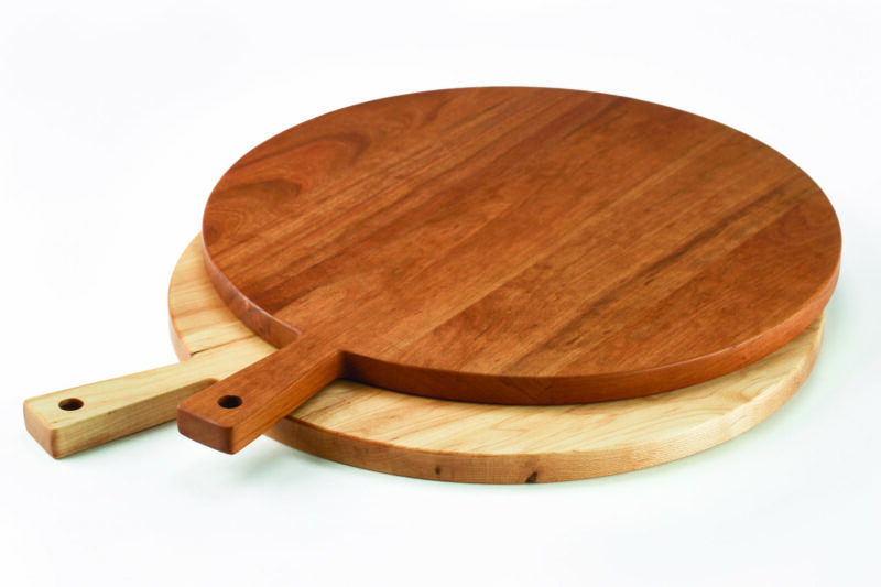 A Maple round wooden serving board lies on top of a Cherry round wooden serving bowl. Each has a small rectangular handle.