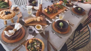 A table set with wooden bowls, plates, serving platters and utensils. There is salad in a bowl, and a cooked chicken on a cutting board.