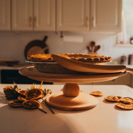 Natural wood cake stand displaying homemade pie in warm kitchen setting with wooden cutting boards and dried citrus decorations